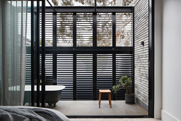 A contemporary bedroom opens onto a private enclosed courtyard or terrace through full-height black-framed glazing. A freestanding white bathtub sits beside the opening, with dark horizontal louvres and screening providing privacy. The space features grey tiled flooring, a dark grey brick feature wall, sheer curtains, a small timber stool, and a potted plant. A portion of the bed is visible in the foreground.