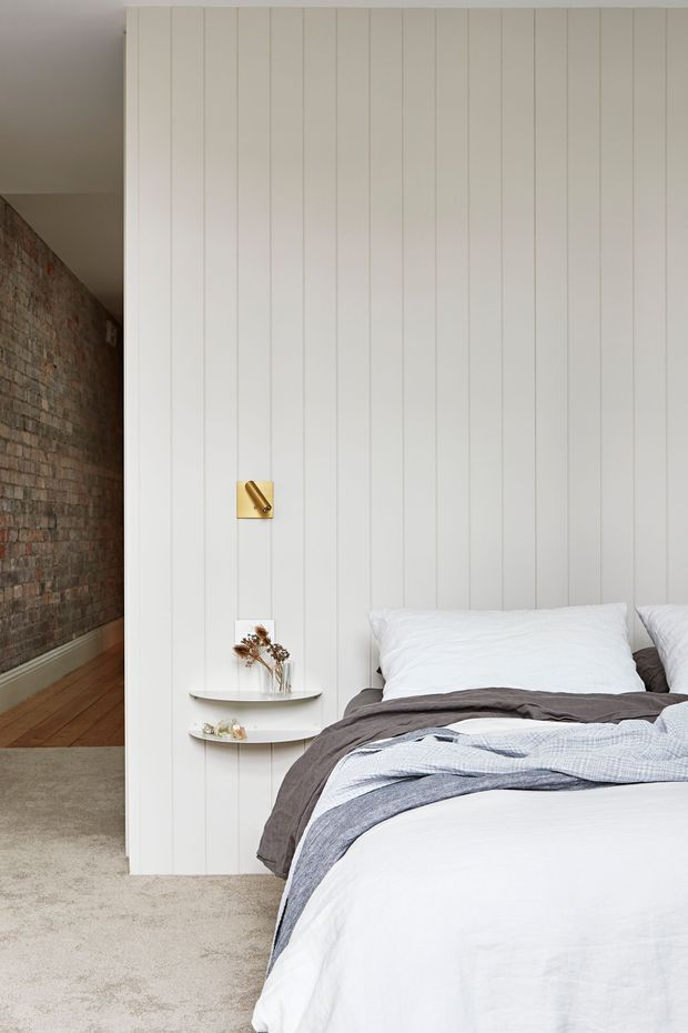 A bright, softly styled bedroom with a white bed positioned against a full-height vertical panelled wall. Layered white, grey and pale blue bedding sits beside small curved floating shelves holding minimal decor, with a compact brass wall light above. A warm exposed brick wall and timber flooring are visible through the opening to the left, while the bedroom floor is carpeted.