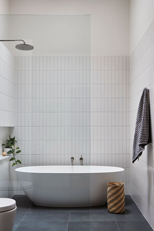 A bright, minimalist bathroom featuring a freestanding white bathtub beneath a large round rainfall showerhead. White vertically arranged wall tiles cover the shower and surrounding walls, while dark grey floor tiles provide contrast. A small plant, woven basket, and patterned towel add natural textural accents.