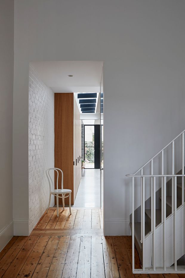 A bright residential hallway and stair landing with original timber floorboards, white-painted brick walls, a white-painted balustrade, and a simple white ceiling. A white bentwood chair sits beside a tall timber built-in cabinet, with a glazed doorway and clerestory-style roof openings visible deeper in the house.
