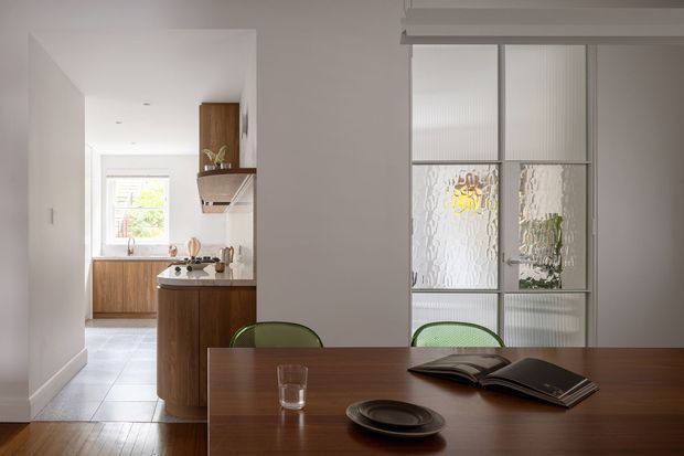 A refined open-plan dining area looking toward a compact kitchen. The foreground features a dark timber dining table with green upholstered chairs, a glass, plate, and open book. Behind it, the kitchen has warm timber cabinetry, a stone benchtop, stainless steel rangehood, and pale tiled flooring. A glazed internal partition with textured translucent glass separates the dining area from another space, while white walls and ceiling create a bright, restrained interior.