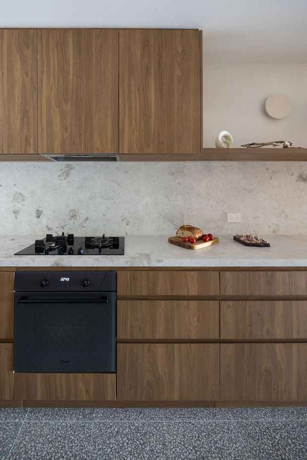 A contemporary kitchen with full-height warm timber cabinetry, a pale textured stone backsplash and benchtop, black integrated oven and gas cooktop, and minimalist open shelving with decorative objects. The lower cabinetry is set above a grey terrazzo-style floor, creating a refined, earthy palette.