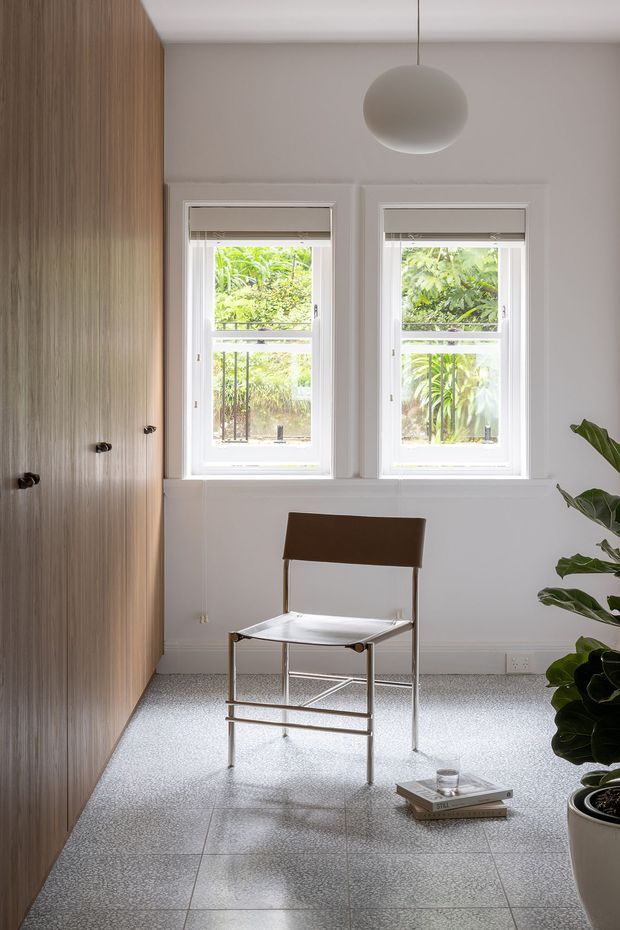 A bright, compact interior with a minimalist workspace or sitting area. The room features full-height vertical timber-grain built-in cabinetry along the left wall, two white-trimmed windows overlooking lush greenery, a simple sculptural pendant light, and a single modern chair positioned on a pale terrazzo-like tiled floor. A large indoor plant occupies the right foreground, adding organic contrast to the restrained palette.