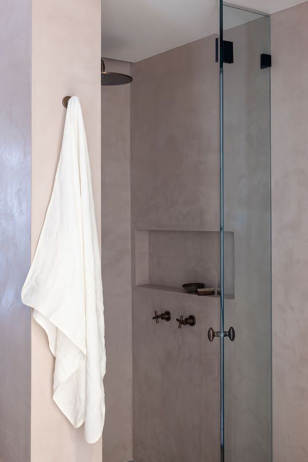 A compact contemporary shower enclosure with frameless glass doors, a large round overhead shower, dark metal controls and hardware, a recessed wall niche, and softly textured taupe plaster-like wall surfaces. A white towel hangs from a circular wall hook in the foreground.