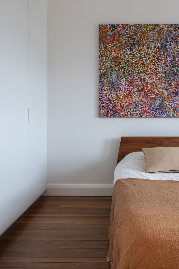 A calm, contemporary bedroom corner with a partially visible bed dressed in a textured warm beige-brown throw and light neutral bedding. A simple timber bedhead and matching dark timber floor contrast with the white walls and built-in white wardrobe. A colourful abstract artwork provides a strong accent above the bed.
