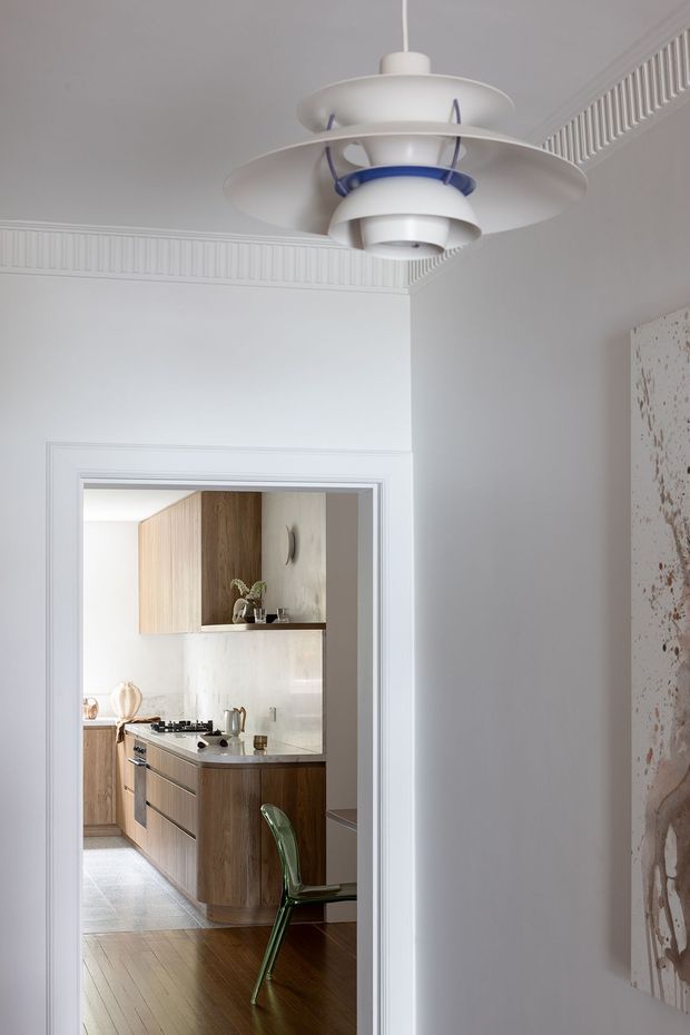 A refined interior view looking through a white-trimmed doorway into a compact contemporary kitchen. The foreground has pale grey-white walls, decorative fluted ceiling trim, a sculptural white pendant light with blue detailing, and a large abstract artwork. The kitchen beyond features warm wood cabinetry, a pale stone benchtop and backsplash, timber flooring, integrated appliances, and a green transparent chair. The space has a restrained, elegant palette with a blend of classic architectural detailing and contemporary furnishings.