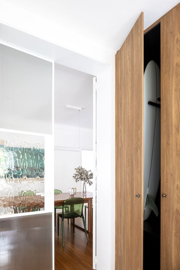 Bright contemporary interior view toward a dining area, framed by white walls and a tall warm timber built-in cupboard. The open cupboard reveals a surfboard or similarly elongated stored object. A translucent ribbed-glass sliding or partition panel with an abstract textured artwork separates the spaces. The dining area contains a timber table, green mesh chairs, a small pendant light, and decorative branches, with polished dark timber flooring.