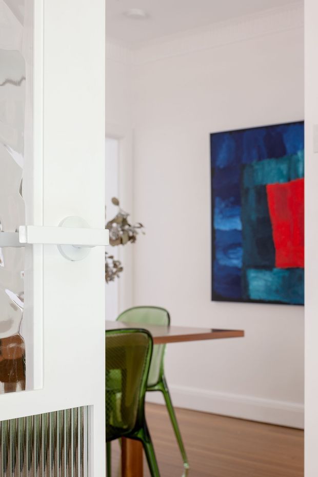 Bright, refined dining area viewed through a partially open white-framed glazed door. The room has white walls with simple trim, warm timber flooring, a timber dining table, translucent green dining chairs, and a large bold abstract artwork in blue, teal, and red. A decorative plant is visible in the background, while the foreground includes a white door handle and ribbed glass panel.