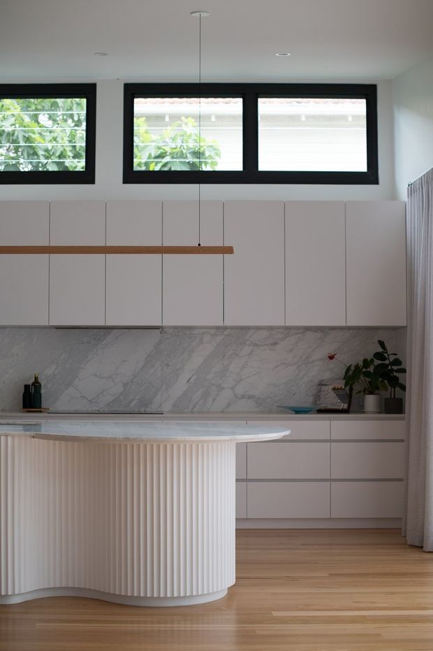 Bright contemporary kitchen featuring a sculptural curved island with ribbed vertical detailing, matte white cabinetry, a dramatic grey-veined marble splashback, pale timber flooring, and black-framed horizontal windows. A slim linear pendant hangs above the island, with soft curtains and indoor plants adding warmth.