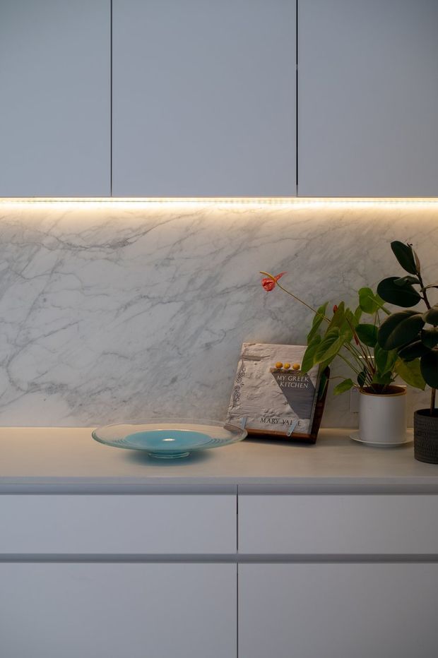 Close-up of a refined modern kitchen built-in, featuring smooth pale grey cabinetry, a white marble-look backsplash with grey veining, and warm linear under-cabinet lighting. A pale countertop displays a shallow glass bowl, cookbook, and potted plants, creating a softly styled, elegant vignette.