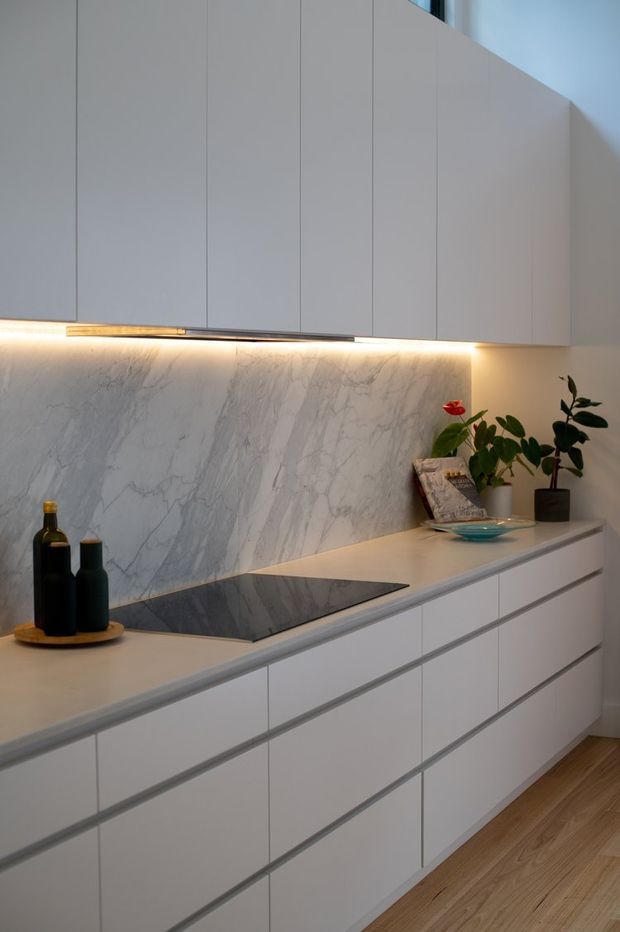 A sleek contemporary kitchen with handleless white cabinetry, a pale stone benchtop and dramatic white marble-look splashback. Warm under-cabinet lighting illuminates the cooking zone, which includes a black glass induction cooktop. Light timber flooring and small potted plants add warmth to the otherwise minimal interior.