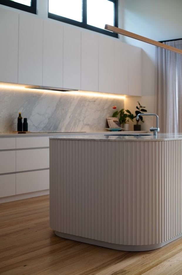 A contemporary kitchen featuring a large rounded island with vertically fluted cabinetry, a pale stone benchtop, white handleless wall and base cabinetry, and a veined stone splashback. Warm under-cabinet lighting, timber flooring, black-framed clerestory windows, and a timber pendant add contrast and warmth.