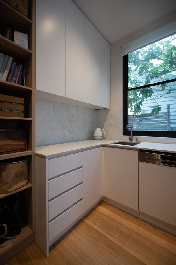 Compact contemporary kitchen with an L-shaped layout, white flat-panel cabinetry, a pale stone benchtop and veined stone splashback. Integrated appliances sit beneath a black-framed window, while open timber shelving provides storage along the left wall. Warm timber flooring contrasts with the soft neutral joinery.