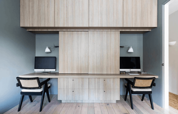 A compact, built-in dual workstation with two computer desks facing each other on either side of a central pale timber storage and shelving unit. Matching black-framed chairs, wall-mounted task lights, blue-grey walls, pale timber cabinetry, and light timber flooring create a clean, contemporary home office. An open doorway is visible at right.