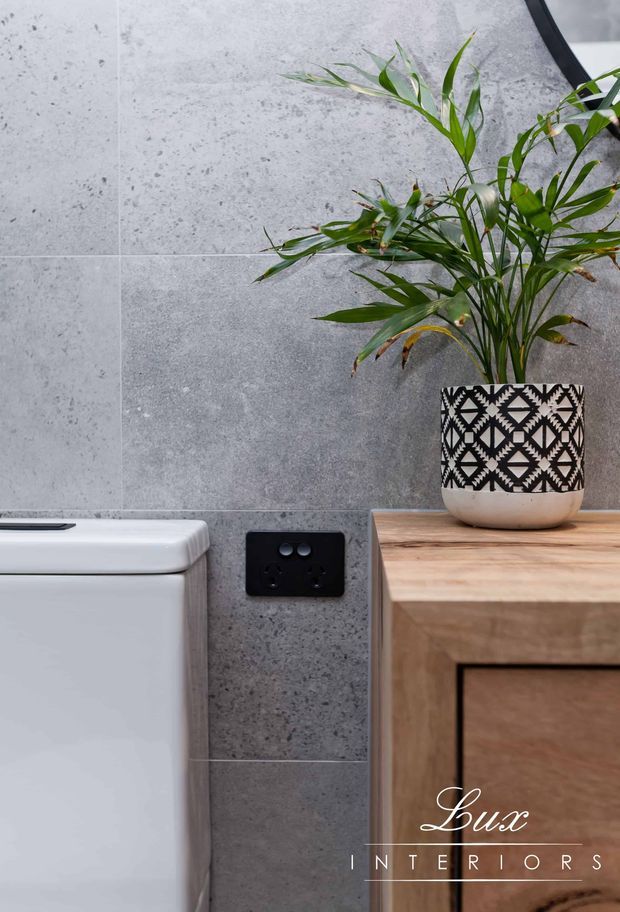 Close-up of a contemporary bathroom interior featuring large-format grey stone-look wall tiles, a white toilet, a black electrical outlet, and a timber vanity or cabinet topped with a geometric black-and-white patterned planter holding a green palm. The image has a clean, modern aesthetic with natural wood and greenery providing warmth.