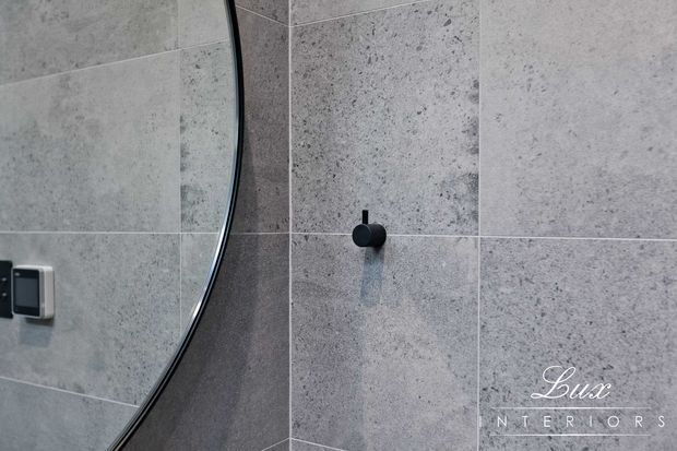 Close-up of a contemporary bathroom wall finished in large-format grey stone-look tiles with fine grout lines. A round-edged mirror, black wall-mounted fixture, and dark switch plate are visible. The image has a cool, refined palette and a Lux Interiors watermark.