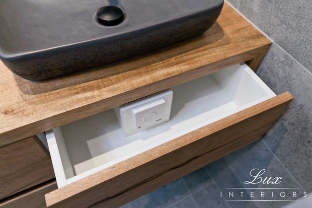 Close-up of a bathroom vanity featuring a dark stone vessel basin on a timber benchtop, with a partially open white-lined drawer and visible power outlet. The vanity has warm wood cabinetry and sits against a grey tiled wall and floor.
