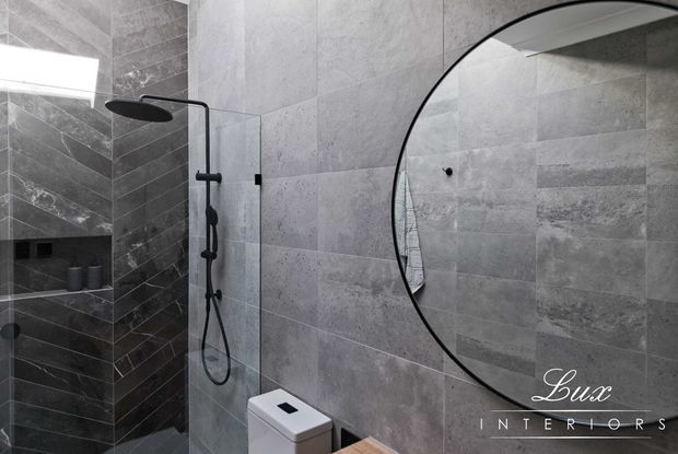 Contemporary bathroom with large-format grey stone-look wall tiles, a darker tiled shower niche and bench, frameless glass shower screen, matte black rain shower and fixtures, a wall-hung toilet, and a large round black-framed mirror. The space has a refined monochrome palette and strong textural contrast.