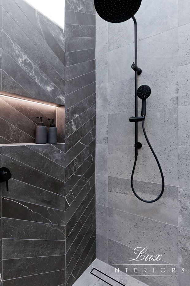 A contemporary walk-in shower with large-format grey stone-look wall tiles, darker veined tiles arranged in a chevron pattern on the recessed shelf wall, and matte black rain shower and handheld fittings. The niche includes integrated warm strip lighting and dark dispensers, while a linear floor drain is visible at the base.