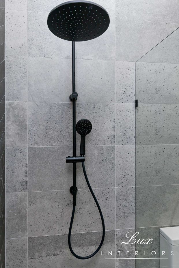 Close-up of a contemporary bathroom shower enclosure featuring a matte black ceiling-mounted rain shower, matching handheld shower, controls, and hose against large-format light grey textured stone-look wall tiles. A frameless glass shower panel and partial toilet are visible at the right, with Lux Interiors branding overlaid near the bottom.