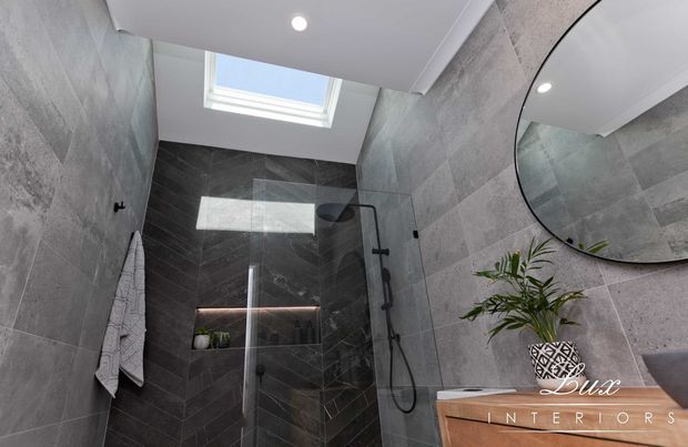 Contemporary bathroom with full-height grey stone-look wall tiling, a dark herringbone-tiled walk-in shower behind clear glass, black shower fittings, a large round wall mirror, timber vanity, recessed shower niche, and a skylight providing natural light.