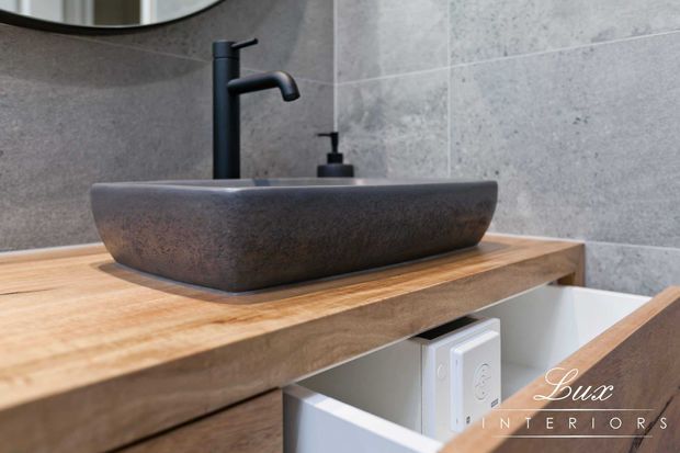 Close-up of a contemporary bathroom vanity featuring a dark stone vessel basin with matte black tapware and soap dispenser, set on a thick timber benchtop with integrated drawer storage. Large-format grey wall tiles and a partial round mirror complete the restrained, tactile scheme.
