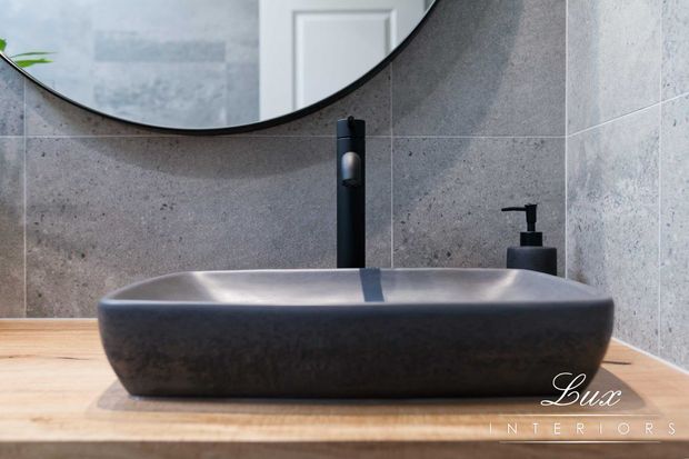 Close-up of a contemporary bathroom vanity featuring a dark rectangular vessel basin on a light timber countertop, a matte black wall-mounted tap and soap dispenser, and large grey stone-look wall tiles. A round mirror is partially visible above the basin.