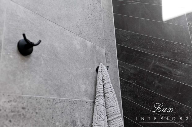 Close-up of a contemporary bathroom wall featuring large-format grey stone-look tiles, a contrasting dark tiled wall, a matte black robe hook, and a textured grey towel. The image has a monochrome, refined interior aesthetic.