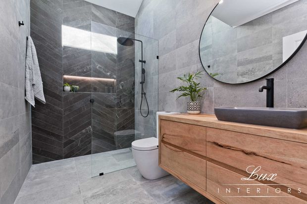 Contemporary bathroom with large-format grey stone-look wall and floor tiles, a frameless glass walk-in shower, wall-hung toilet, and a timber vanity with a dark vessel basin and matte black tapware. A large round mirror and concealed warm lighting add contrast and refinement.