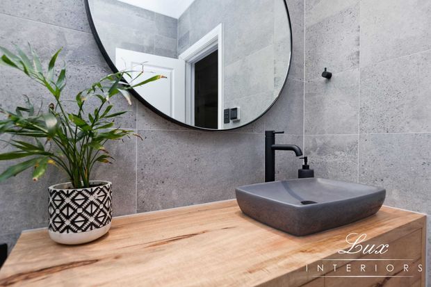 Contemporary bathroom vanity featuring large-format grey stone-look wall tiles, a thick natural timber benchtop, a dark vessel basin with matching matte-black tapware, and a round black-framed mirror. A patterned planter adds a decorative accent.
