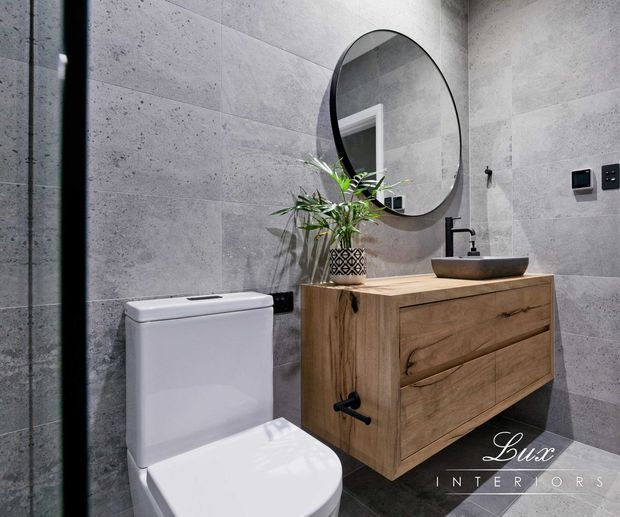 Contemporary bathroom featuring large grey concrete-look wall tiles, a wall-mounted timber vanity with integrated drawers, a vessel basin and matte black tapware. A round black-framed mirror, white toilet and potted greenery complete the refined, industrial-inspired interior.