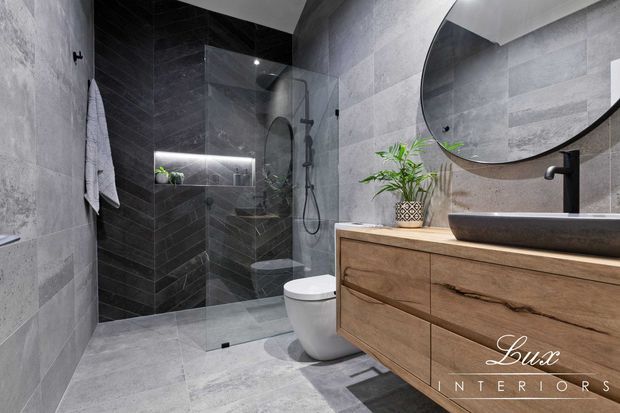 Contemporary bathroom featuring large-format grey stone-look wall and floor tiles, a dark tiled walk-in shower with clear glass screen, wall-hung toilet, and a floating natural timber vanity with a vessel basin and matte black tapware. A large round mirror and subtle integrated shower lighting complete the refined, monochromatic scheme.
