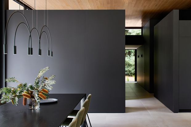 A dramatic contemporary dining area with a long dark dining table, sculptural suspended pendant lights, and textured upholstered chairs. A dark charcoal partition wall separates the dining space from a minimalist hallway, while warm timber ceiling boards and pale flooring create contrast. Full-height glazing and views of greenery are visible beyond the corridor.