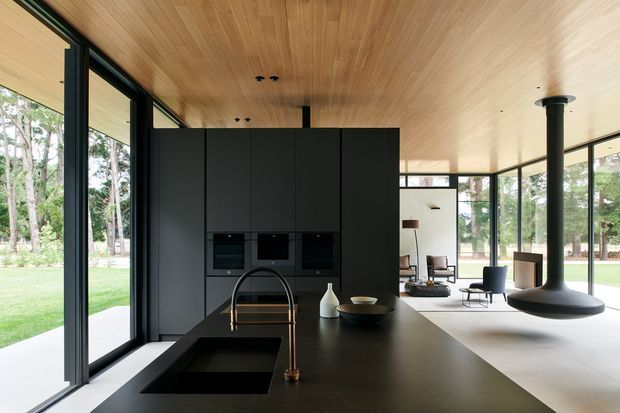 Open-plan contemporary kitchen and living area with a large dark island, integrated black cabinetry and appliances, timber-lined ceiling, extensive black-framed glazing, and a sculptural suspended fireplace. The space opens directly onto a landscaped garden and has a calm, refined indoor-outdoor character.