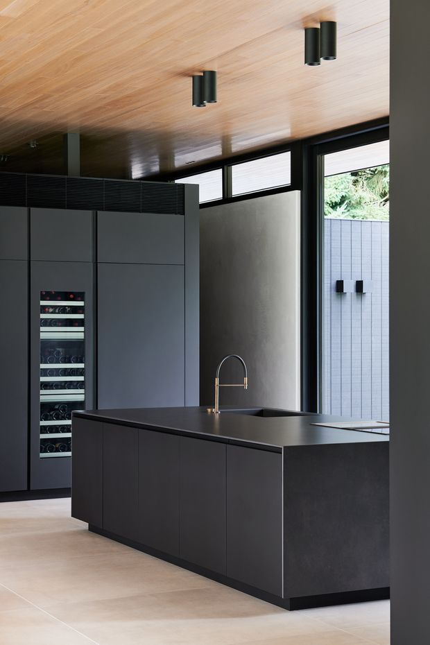 A sophisticated contemporary kitchen with a large dark charcoal island, integrated sink and brass-toned faucet, tall dark cabinetry, and a built-in wine refrigerator. The room features a warm timber-lined ceiling, pale large-format flooring, dark-framed glazing, and a textured light wall. The restrained palette and clean geometric joinery create a refined, modern atmosphere.