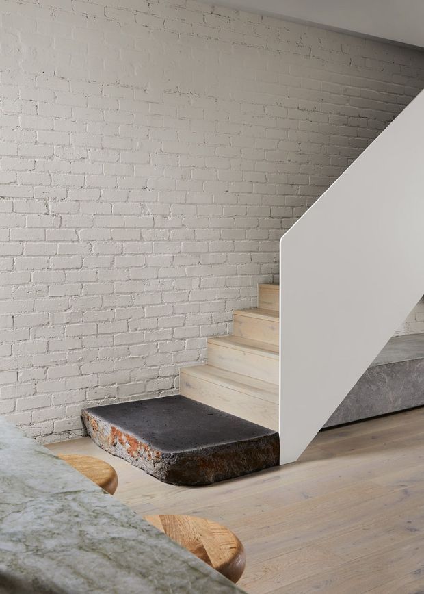 Interior architectural detail showing a minimalist staircase with pale timber treads, a sculptural white side wall and handrail-free balustrade, and a substantial dark stone slab forming the lower landing. The background wall is white-painted exposed brick, while pale timber flooring and part of a mottled stone countertop or table are visible in the foreground. The composition combines clean contemporary geometry with raw industrial materials.