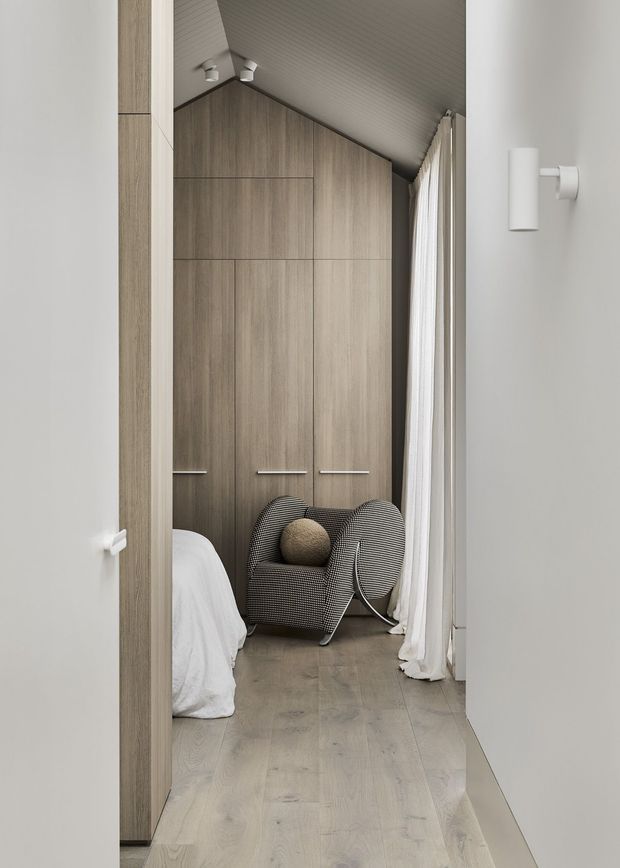 A narrow, refined bedroom viewed through an open doorway, featuring full-height timber wardrobe cabinetry, pale timber flooring, a sloped ribbed ceiling, sheer full-height curtains, and a sculptural upholstered lounge chair with a round cushion. The palette is soft grey, warm light wood, and off-white, creating a calm contemporary interior.