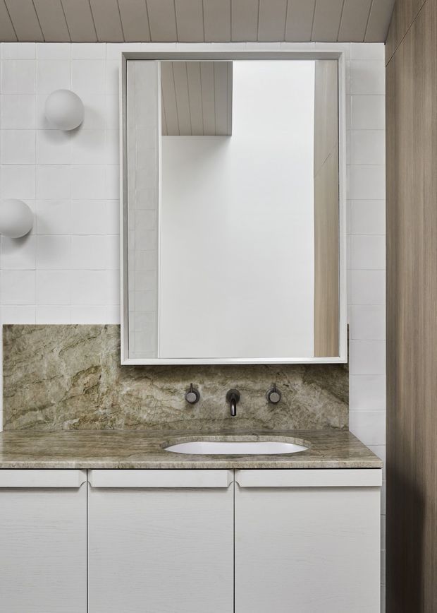 A compact contemporary bathroom vanity featuring a white inset basin, wall-mounted dark metal tapware, and a natural stone benchtop with matching full-height splashback. White rectangular wall tiles, pale timber-look cabinetry, a large framed mirror, and a light painted timber-look ceiling create a clean, understated finish.