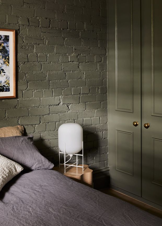 A moody bedroom interior featuring a bed with muted mauve-grey bedding, a dark olive-grey exposed brick feature wall, and coordinated paneled built-in wardrobe doors with brass knobs. A small timber bedside table holds a sculptural white lamp, while framed artwork is partially visible on the wall.
