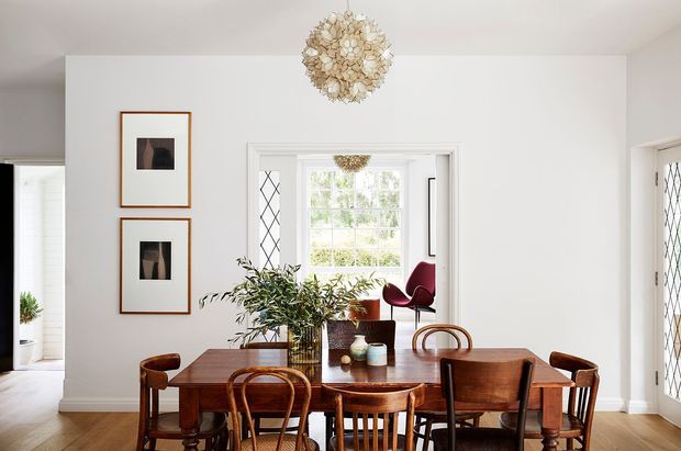 Bright traditional dining room with a polished dark timber dining table surrounded by mixed timber chairs. White walls, decorative diamond-leaded glazing, framed artwork, indoor plants, and a sculptural floral pendant light create a refined, warm interior. A red lounge chair is visible through the adjoining opening.