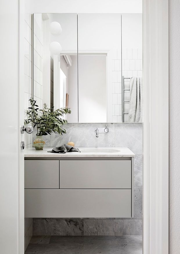 Bright, compact contemporary bathroom viewed through a doorway, featuring a floating grey vanity with integrated basin, pale marble-look wall and floor surfaces, large frameless mirrored cabinetry, white subway wall tiles, chrome tapware, and a ladder towel rail. A small plant and neutral textiles add softness to the otherwise clean, minimal scheme.