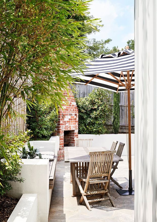 Bright landscaped courtyard patio viewed from beside a white-framed opening, with a timber dining table and folding chairs beneath a large striped cantilever-style umbrella. White rendered planter walls, a red brick outdoor fireplace, timber fencing, lush bamboo and shrubs create a private garden setting.