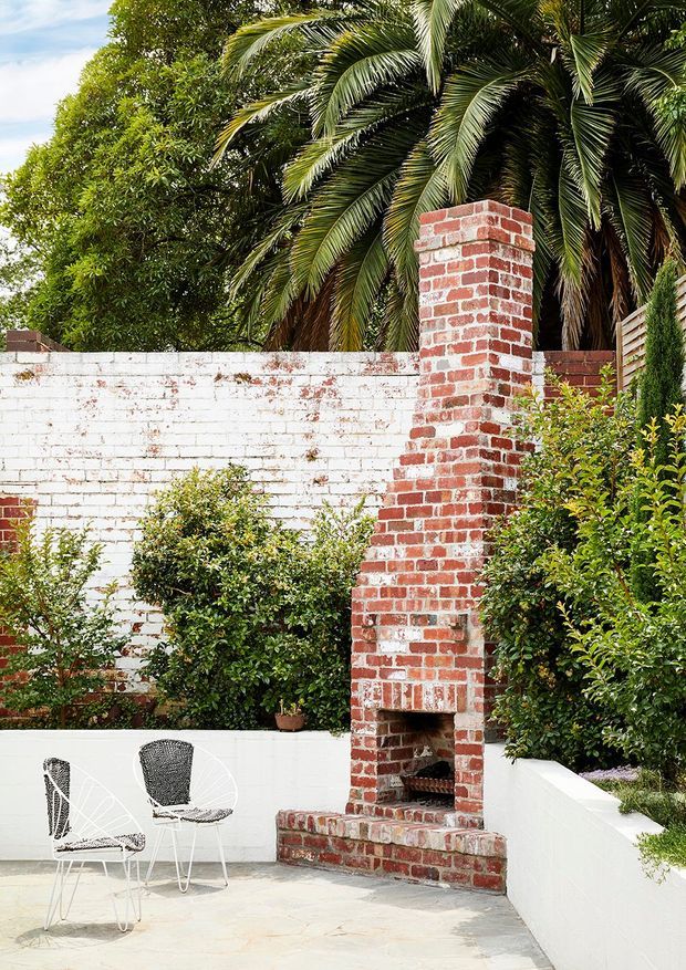 A bright landscaped courtyard or patio with a tall exposed red-brick outdoor fireplace set into a white-painted brick wall. White rendered masonry planter walls, clipped shrubs, palm foliage, timber screening, and two lightweight wire-frame chairs create a relaxed garden entertaining area.