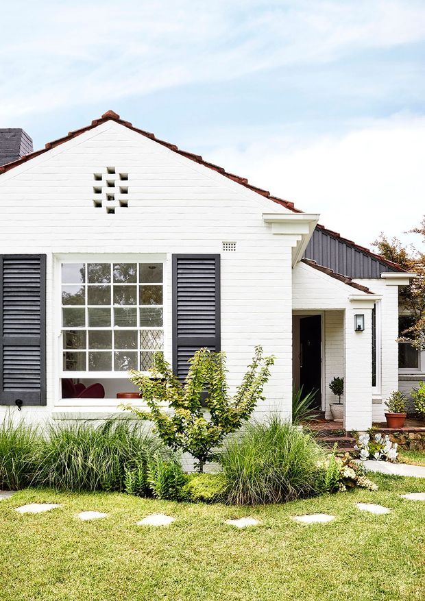 Front exterior view of a compact white-painted brick and weatherboard-style house with a gabled terracotta-tiled roof, dark louvred shutters, multi-pane windows, and a small covered entry. A landscaped front garden with ornamental grasses, shrubs, stepping stones, and lawn occupies the foreground.
