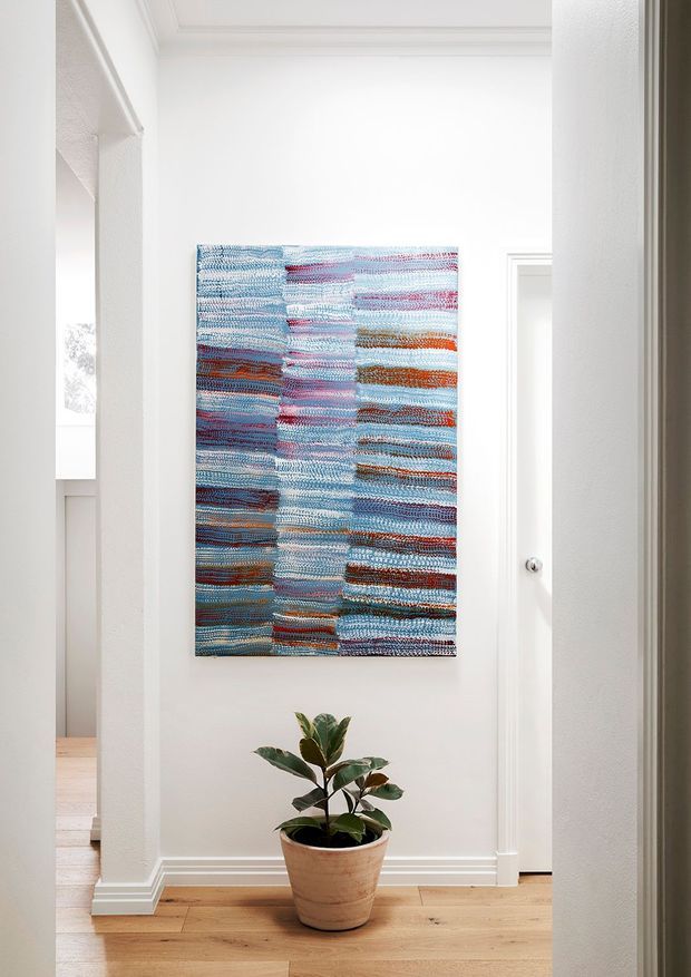 Bright, narrow residential hallway with white painted walls, decorative ceiling moulding, white door and trim, and a warm timber floor. A colourful woven abstract textile artwork is mounted on the feature wall above a potted indoor plant. An adjoining room is partially visible through the opening on the left.
