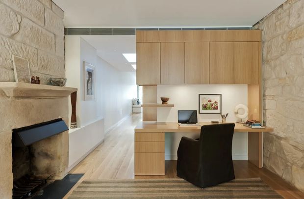 A warm, compact home office integrated into a timber built-in joinery wall. The desk includes open shelving, drawers, overhead cupboards, a laptop and accessories, with a dark upholstered chair positioned on a striped rug. Rough exposed stone walls and a substantial stone fireplace frame the room, while pale timber flooring and a white corridor create a bright, refined setting.