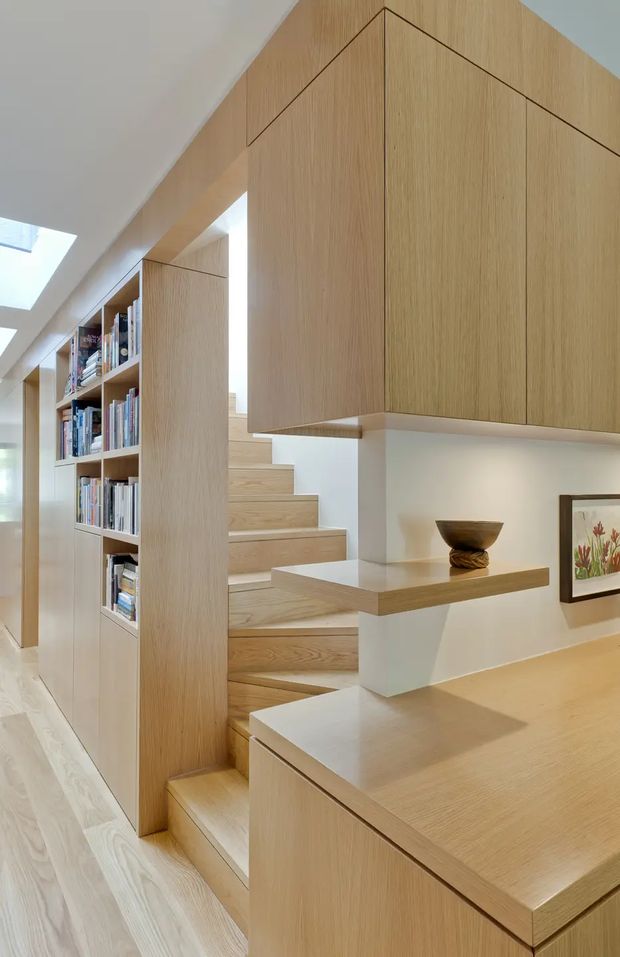 Bright contemporary interior showing a compact timber-lined stairway integrated with extensive built-in cabinetry and open bookshelves. Pale oak-toned joinery wraps around the stair, with a floating display shelf, light timber flooring, white walls, skylights, and framed artwork. The design is highly tailored and space-efficient, with clean flush doors and concealed storage.