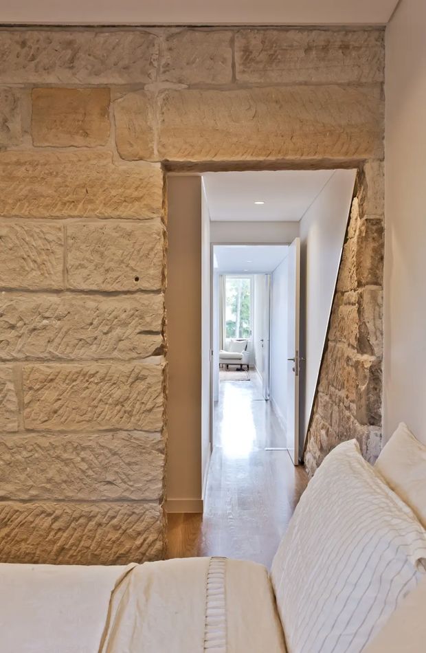 A bedroom viewed toward a bright hallway, featuring a prominent exposed textured stone wall and stone-framed doorway. The bed has white bedding with subtle linear detailing, while the hallway has pale walls, light timber flooring, and recessed ceiling lighting, leading toward a softly furnished room and window.