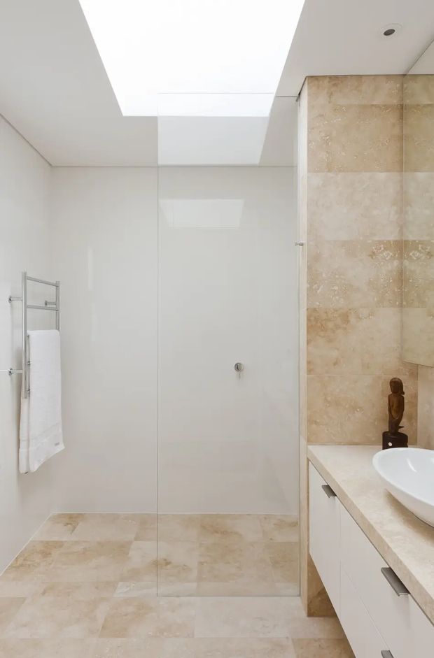 Bright minimalist bathroom featuring a frameless glass walk-in shower, large beige natural-stone tiles on the floor and shower wall, white wall surfaces, a skylight, a wall-mounted towel rail, and a pale stone vanity with white built-in cabinetry and basin.