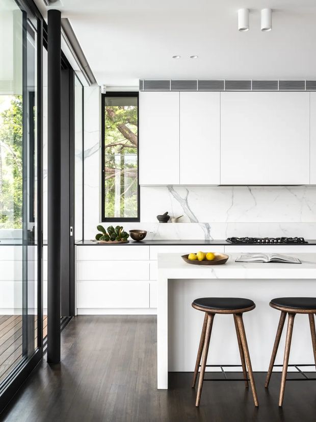 Bright contemporary kitchen with a white marble-look splashback and benchtops, streamlined white overhead and lower cabinetry, black-framed floor-to-ceiling glazing, dark timber flooring, and a central island with two dark timber stools. Minimal detailing and integrated ventilation create a clean, refined interior.